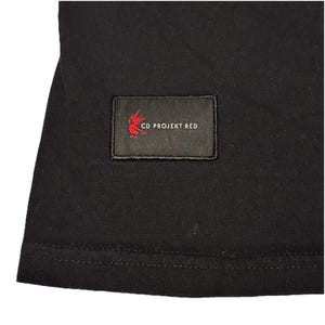 May include: Black t-shirt with a rectangular patch featuring the text "CD PROJEKT RED" in white and a red stylized design. The patch is sewn onto the shirt. The shirt is made of a dark fabric.