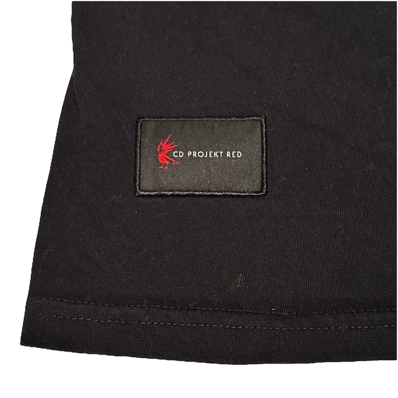 May include: Black t-shirt with a rectangular patch featuring the text "CD PROJEKT RED" in white and a red stylized design. The patch is sewn onto the shirt. The shirt is made of a dark fabric.