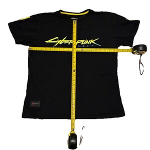 May include: Black t-shirt with the text "Cyberpunk 2077" in neon yellow and blue. The shirt has a yellow collar and a small patch on the lower left side. A yellow measuring tape is laid across the shirt.