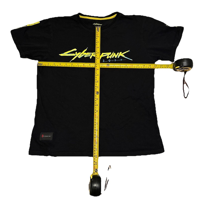 May include: Black t-shirt with the text "Cyberpunk 2077" in neon yellow and blue. The shirt has a yellow collar and a small patch on the lower left side. A yellow measuring tape is laid across the shirt.