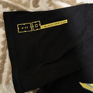 May include: Black t-shirt with yellow graphics. The design includes a series of arrows and a geometric shape, along with the text "MANTIS BLADE SYSTEM MOD 3". The shirt is made of a soft, dark fabric.
