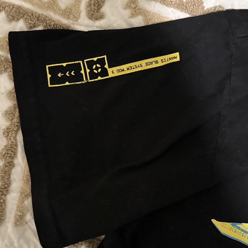 May include: Black t-shirt with yellow graphics. The design includes a series of arrows and a geometric shape, along with the text "MANTIS BLADE SYSTEM MOD 3". The shirt is made of a soft, dark fabric.