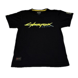 May include: Black t-shirt with the text "Cyberpunk 2077" in yellow and blue. The shirt has a yellow collar and a small logo on the sleeve. A small red tag is visible on the bottom left.