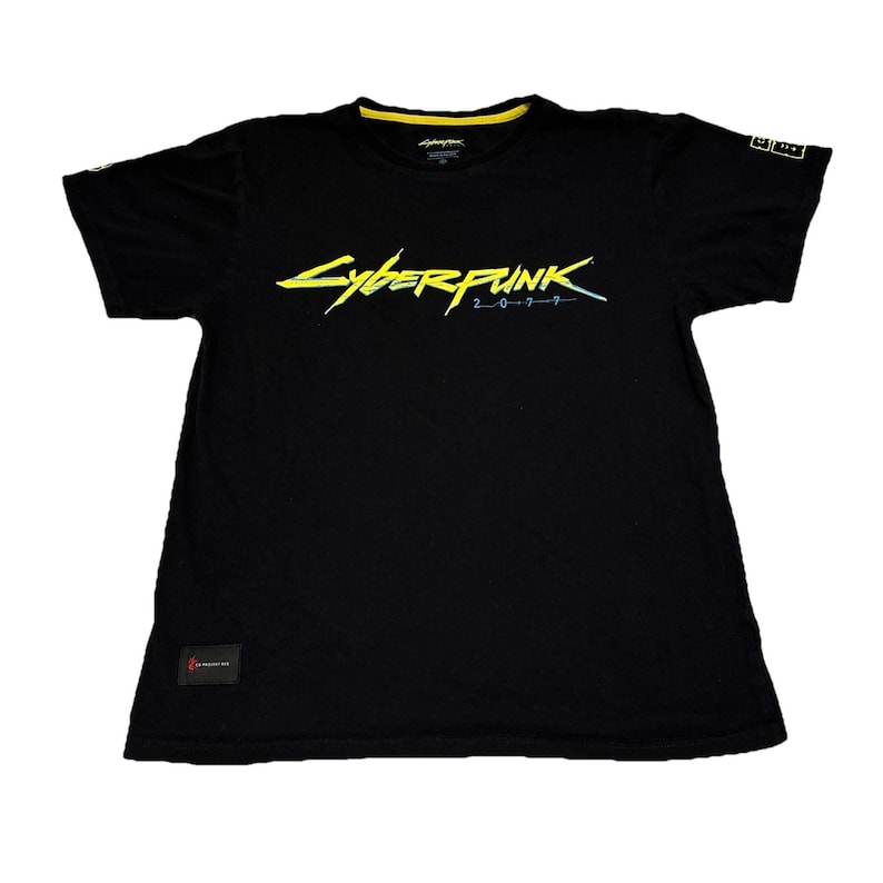 May include: Black t-shirt with the text "Cyberpunk 2077" in yellow and blue. The shirt has a yellow collar and a small logo on the sleeve. A small red tag is visible on the bottom left.
