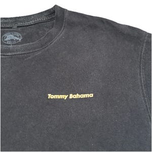 May include: Black crew neck sweatshirt with the embroidered text "Tommy Bahama" in gold. The top has a small logo near the neckline. The sweatshirt is made of a soft material.