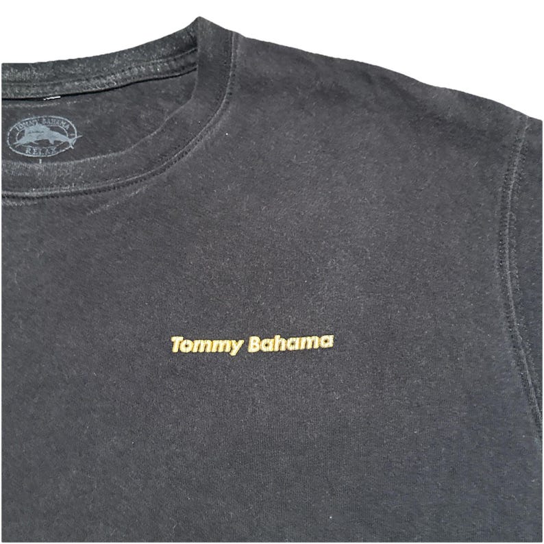 May include: Black crew neck sweatshirt with the embroidered text "Tommy Bahama" in gold. The top has a small logo near the neckline. The sweatshirt is made of a soft material.