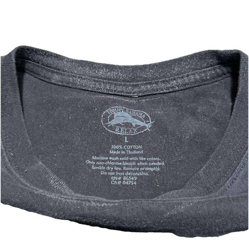 May include: Close-up of a black Tommy Bahama Relax t-shirt tag. The tag includes the brand logo, size 'L', and care instructions. The shirt is made of 100% cotton and made in Thailand.