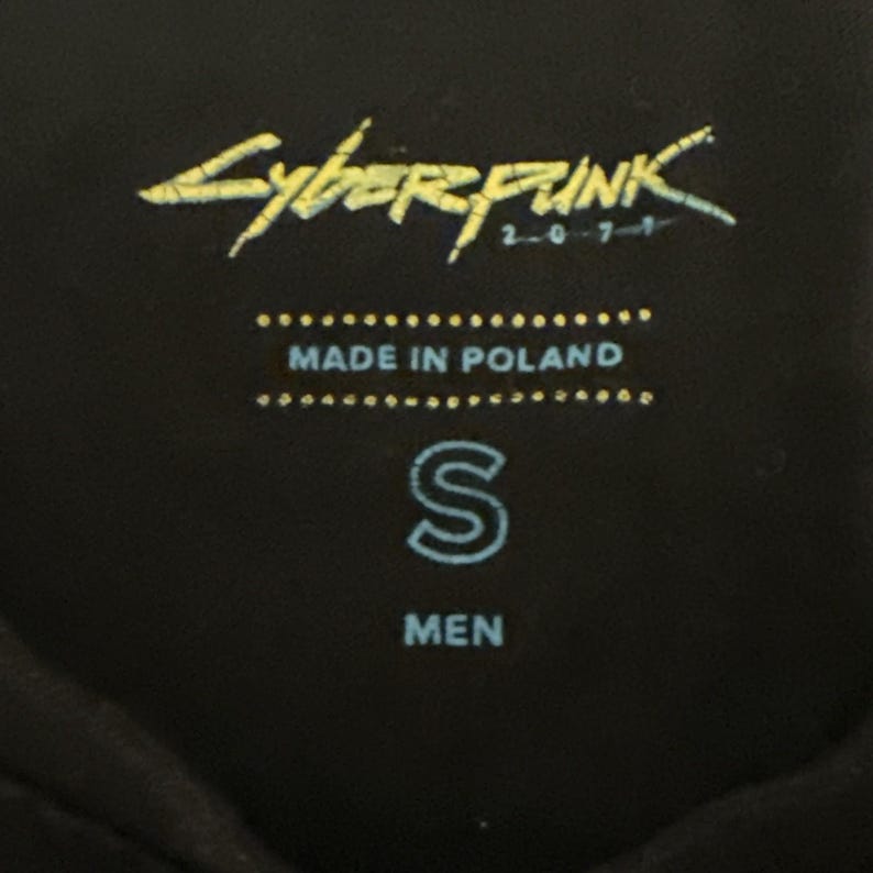 May include: Black garment tag with the text "Cyberpunk 2077" in yellow. Below the text are the words "MADE IN POLAND" and the letter "S". The word "MEN" is printed below the letter "S".