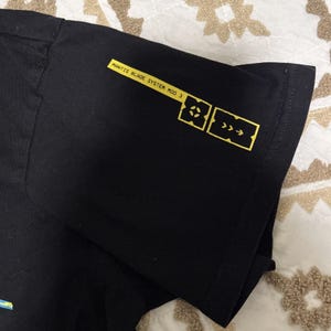 May include: Black t-shirt with yellow text and graphics on the sleeve. The text reads "MANTIS BLADE SYSTEM MOD 3". The graphics include a square with a cross and a square with arrows.
