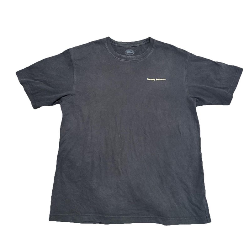May include: A dark gray t-shirt with the Tommy Bahama logo on the chest. The shirt has a crew neck and short sleeves. The fabric appears to be a soft, worn cotton blend. The shirt is laid flat on a white surface.