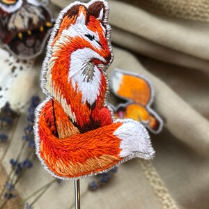 Pode incluir: Um pequeno broche de raposa bordado com pele laranja e branca, sentado em um alfinete de metal. A raposa está olhando para a direita e tem a cauda enrolada ao redor do seu corpo.