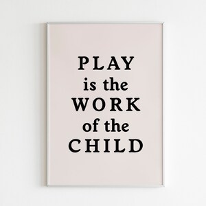 Può includere: Una stampa incorniciata con uno sfondo rosa chiaro e testo nero che recita "PLAY is the WORK of the CHILD."