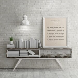 May include: A white framed print of a book excerpt from "The Sun Also Rises" by Ernest Hemingway. The print is hanging on a white brick wall above a whitewashed wooden shelf with a stack of books. A small potted plant sits on the shelf.