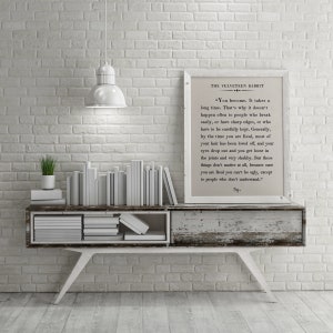 May include: A white brick wall with a white shelf and a framed print of a passage from "The Velveteen Rabbit" by Margery Williams. The print is on a white background with black text. The shelf is decorated with a stack of white books and a small green plant in a white pot.