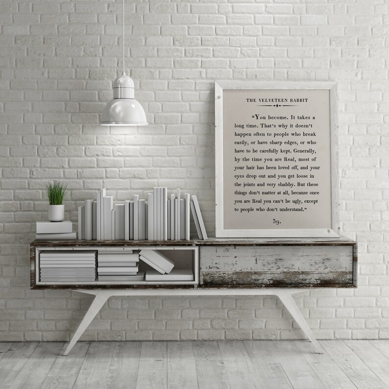 May include: A white brick wall with a white shelf and a framed print of a passage from "The Velveteen Rabbit" by Margery Williams. The print is on a white background with black text. The shelf is decorated with a stack of white books and a small green plant in a white pot.