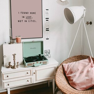 May include: A light pink framed print with the text "I found home in love, not in cities."  A green vintage record player sits on a white dresser with three drawers. A wicker chair with a pink cushion sits in the corner of the room.