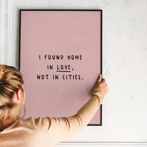 May include: A person hangs a pink framed print on a white wall. The print features black text that reads "I found home in love, not in cities."