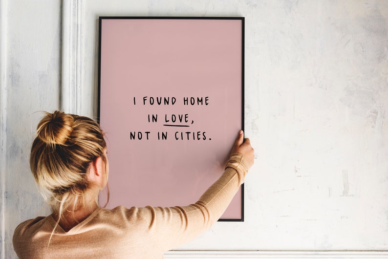 May include: A person hangs a pink framed print on a white wall. The print features black text that reads "I found home in love, not in cities."