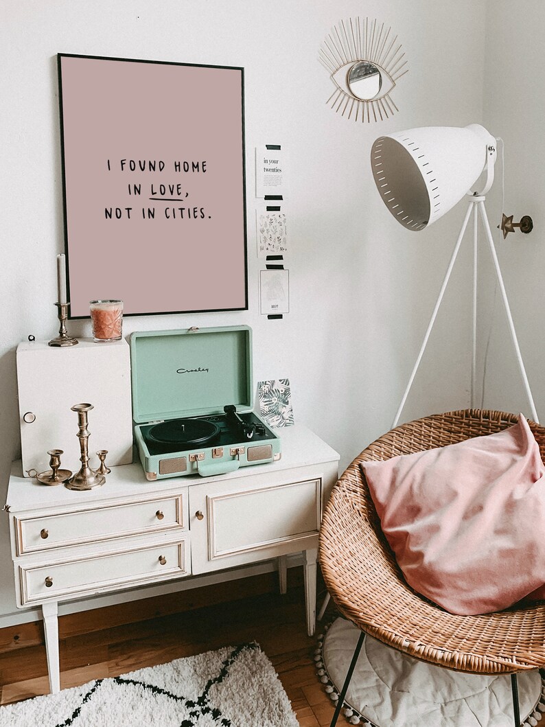 May include: A light pink framed print with the text "I found home in love, not in cities."  A green vintage record player sits on a white dresser with three drawers. A wicker chair with a pink cushion sits in the corner of the room.