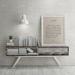 K&ouml;nnte beinhalten: Eine wei&szlig;e Backsteinwand mit einer wei&szlig;en Lampe, die von der Decke h&auml;ngt. Ein wei&szlig;es Regal mit einem Stapel wei&szlig;er B&uuml;cher und einer kleinen gr&uuml;nen Pflanze steht auf dem Regal. Ein gerahmter Druck mit einem Zitat von A.A. Milne h&auml;ngt an der Wand. Das Zitat lautet: "Wenn es jemals ein Morgen gibt, an dem wir nicht zusammen sind, dann gibt es etwas, das du immer im Ged&auml;chtnis behalten solltest. Du bist mutiger, als du glaubst, st&auml;rker, als du scheinst, und kl&uuml;ger, als du denkst. Aber das Wichtigste ist, selbst wenn wir getrennt sind, werde ich immer bei dir sein."