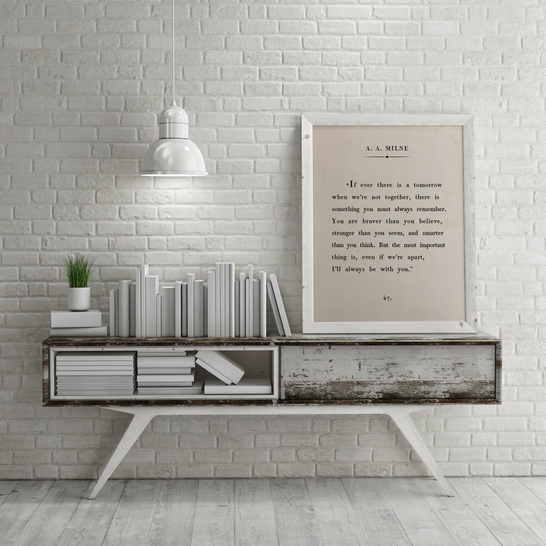 K&ouml;nnte beinhalten: Eine wei&szlig;e Backsteinwand mit einer wei&szlig;en Lampe, die von der Decke h&auml;ngt. Ein wei&szlig;es Regal mit einem Stapel wei&szlig;er B&uuml;cher und einer kleinen gr&uuml;nen Pflanze steht auf dem Regal. Ein gerahmter Druck mit einem Zitat von A.A. Milne h&auml;ngt an der Wand. Das Zitat lautet: "Wenn es jemals ein Morgen gibt, an dem wir nicht zusammen sind, dann gibt es etwas, das du immer im Ged&auml;chtnis behalten solltest. Du bist mutiger, als du glaubst, st&auml;rker, als du scheinst, und kl&uuml;ger, als du denkst. Aber das Wichtigste ist, selbst wenn wir getrennt sind, werde ich immer bei dir sein."