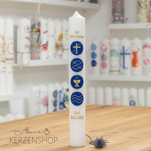 Könnte beinhalten: Eine weiße Kommunionkerze mit blauen und goldenen religiösen Symbolen und Text. Die Kerze hat ein Kreuz, Fische, einen Kelch und Wellen. Der Text lautet "Zur Kommunion" und "Carl 12.12.2023". Weitere Kerzen sind im Hintergrund zu sehen.