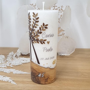 Könnte beinhalten: Eine weiße Stumpenkerze mit einem Baumdesign und den Namen "Carina" und "Paolo". Die Kerze hat einen Holzmaserungsfuß und das Datum "17. Juli 2026" mit Eheringen. Ein dekorativer Artikel für Hochzeiten oder besondere Anlässe.