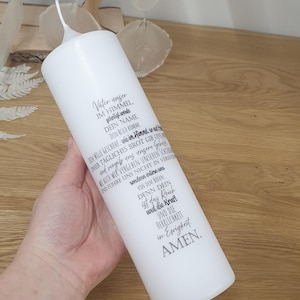 Könnte beinhalten: Eine weiße Säulenkerze mit schwarzem Text. Der Text ist ein Gebet auf Deutsch. Die Kerze ist zylindrisch mit einer leicht gebogenen Oberseite. Die Kerze wird in einer Hand gehalten, der Hintergrund ist eine Holzoberfläche.
