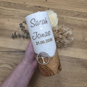 Könnte beinhalten: Eine zylindrische Kerze mit einem weißen, strukturierten oberen Teil und einem unteren Teil mit natürlicher Holzmaserung. Der weiße Bereich zeigt die Namen Sarah und Jonas mit einem kleinen Herz und dem Datum 21.06.2026. Ein runder, hölzerner Lebensbaum-Anhänger ist mit einer Schnur in der Mitte befestigt.