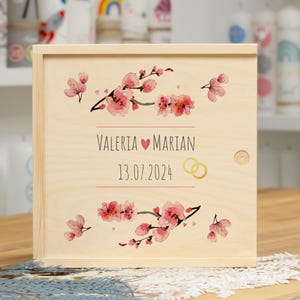 Könnte beinhalten: Eine Holzkiste mit hellem Holzfinish, verziert mit rosa floralen Designs und den Namen "Valeria" und "Marian" mit einem Herzsymbol. Das Datum "13.07.2024" und goldene Eheringe sind ebenfalls abgebildet.