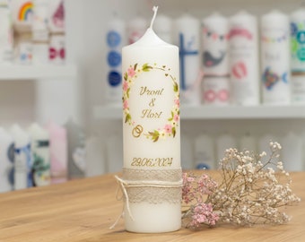 Puede incluir: Vela de boda blanca con un diseño de corona floral y los nombres "Vront & Flort" y la fecha "29.06.2024" escritos en oro. La vela está decorada con una cinta de arpillera.