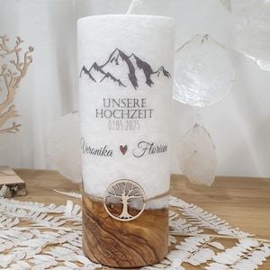 Könnte beinhalten: Weiße Stumpenkerze mit einem Bergpanorama-Design und dem aufgedruckten Text "UNSERE HOCHZEIT 07.05.2025 Veronika Florian". Die Kerze steht auf einem Holzsockel mit einem Baum-Ausschnitt.