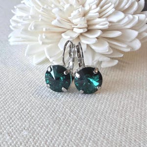 May include: A pair of leverback earrings featuring round, faceted, emerald green crystals set in silver-toned metal. The earrings are displayed against a soft, white background with a large, white flower in the background.