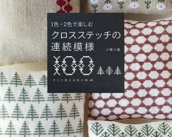 May include: A book cover with the title "100 Cross Stitch Patterns" in Japanese, surrounded by four embroidered pillows. The pillows feature various cross-stitch designs in red, white, and green, including geometric patterns and tree motifs. The book is black.
