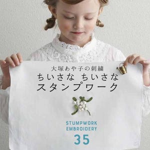 以下が含まれることがあります： 日本語のテキスト、英語の「STUMPWORK EMBROIDERY」、数字の35、白い実と緑の葉の小さな刺繍が施された白い布。右上隅には小さな金属製のじょうろが取り付けられています。