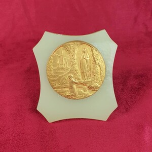Peut inclure: Une plaque blanche avec un médaillon rond de couleur or représentant une image religieuse de la Vierge Marie et une figure agenouillée. Le médaillon est entouré d'une bordure décorative.