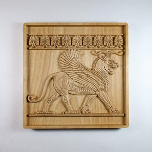 May include: A square light-colored wooden wall plaque featuring a detailed relief carving of a winged mythological creature with horns. The design includes a horizontal decorative border of repeating fan-shaped motifs above the central figure.