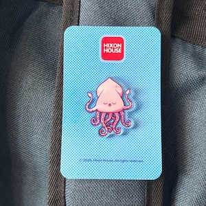 Puede incluir: Un pin rectangular azul claro con un diseño de calamar rosa de dibujos animados. El pin tiene el texto "Hixon House" en un cuadrado rojo en la parte superior. El fondo es un patrón de puntos azules. El pin está sujeto a una mochila azul y gris.