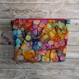 May include: A colorful zippered pouch with a vibrant abstract pattern in shades of blue, yellow, pink, and purple. The pouch has a magenta zipper and a small charm. The bag is sitting on a wooden surface.