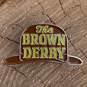 Puede incluir: Un pin de esmalte marrón y amarillo con forma de sombrero con el texto "The Brown Derby" en él.