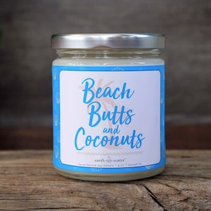 May include: A white soy candle in a glass jar with a blue label that reads "Beach Butts and Coconuts" by "earth & water". The candle is on a wooden surface.