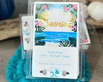 May include: A white wax melt label with a tropical sunrise scene and the text "Sunrise" in a gold font. The label also includes the text "Tropical Mango, Citrus, Star Apple, Papaya" and "Hand-Poured Soy Melts 5.5 oz. Houston TX". The label is surrounded by a clear plastic clamshell.