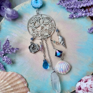 May include: A silver dreamcatcher with a seashell design, featuring a clear quartz crystal, iridescent seashells, and blue and purple crystals hanging from it.