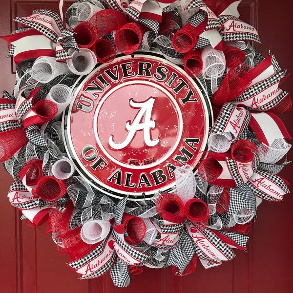 May include: A red, white, and black wreath with a University of Alabama sign in the center. The sign is red with a white outline and the words "University of Alabama" in white. The letter "A" is in the center of the sign and is white with a red outline. The wreath is made of red and white mesh and has black and white checkered ribbon.