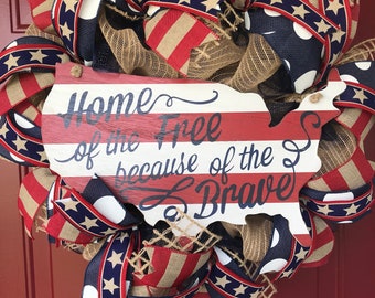 May include: A patriotic wreath featuring red, white, and blue ribbon, burlap, and mesh. The wreath is decorated with a wooden sign that reads "Home of the Free because of the Brave".