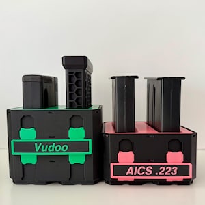 May include: Two black and colored storage containers. One container has green accents and the word "Vudoo" on a green label. The other container has pink accents and the text "AICS .223" on a pink label. Both containers have black tops.