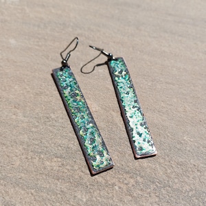 Può includere: Un paio di orecchini pendenti rettangolari. Gli orecchini hanno una cornice color rame con un design verde e argento maculato e strutturato. I ganci degli orecchini sono neri. Gli orecchini sono esposti su una superficie dai toni neutri.