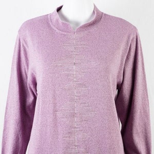 Könnte beinhalten: Ein fliederfarbener Pullover mit langen Ärmeln und einem Mock-Neck-Ausschnitt. Der Pullover hat einen strukturierten Strickstoff und ein vertikales, rautenförmiges Design in einem helleren Farbton in der Mitte. Die Ärmel haben eine leichte Puffärmelform.
