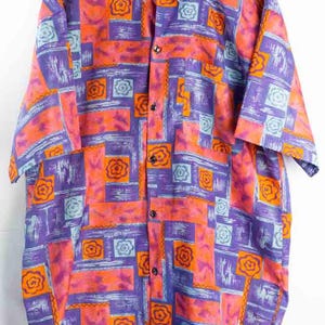 Könnte beinhalten: Kurzärmeliges Button-Down-Hemd mit einem lebendigen geometrischen Muster. Das Hemd hat ein sich wiederholendes Design aus orangefarbenen, lila und blauen Formen, einschließlich floraler und quadratischer Elemente. Es hat einen Kragen und eine Brusttasche.