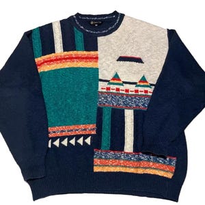 May include: A navy blue sweater with a colour block design. The sweater features sections of teal, cream, and orange, with geometric patterns. The sleeves are solid navy blue. The sweater has a crew neck.